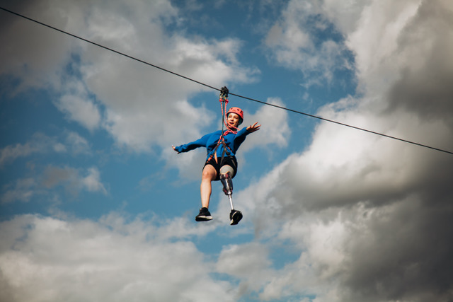 A camper with a prosthetic leg flying down a zipline against a bright sky