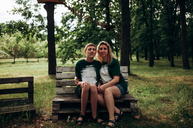 A camper and a staff member sitting side by side on a wooden bench in the woods